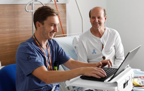 Patient i sjukhussäng och personal vid dator bredvid sängen.