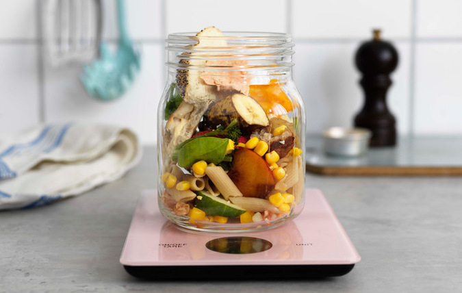 Matrester i from av banan, majs,bröd,fisk, gurka, köttbulle,sallad som nästan helt fyller genomskinlig glasburk som placerats på en digital rosa köksvåg.