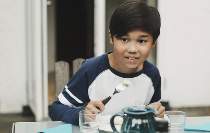 Pojke vid lunchbord med kniv och gaffel i handen tittar på flicka i förgrunden.