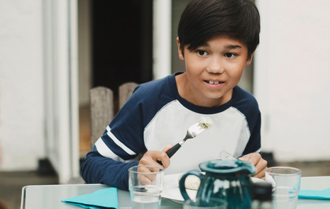 Pojke vid lunchbord med kniv och gaffel i handen tittar på flicka i förgrunden.