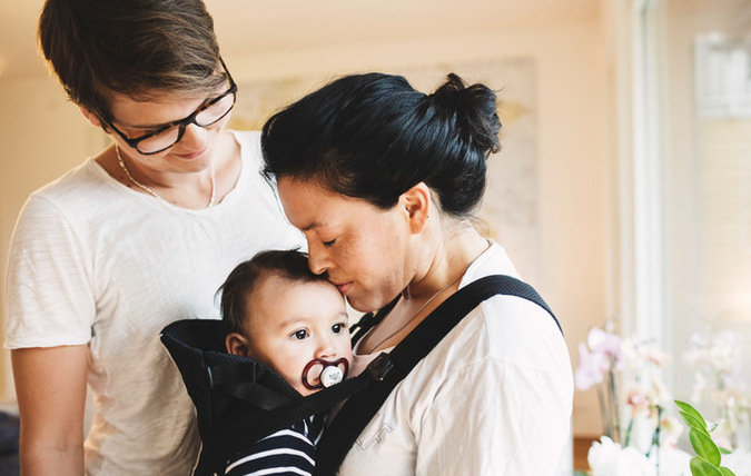 En vuxen person med ett barn i en sele på magen. Hon har munnen mot barnets panna. Bredvid står en annan person och tittar glatt på dem. Barnet har en napp i munnen.